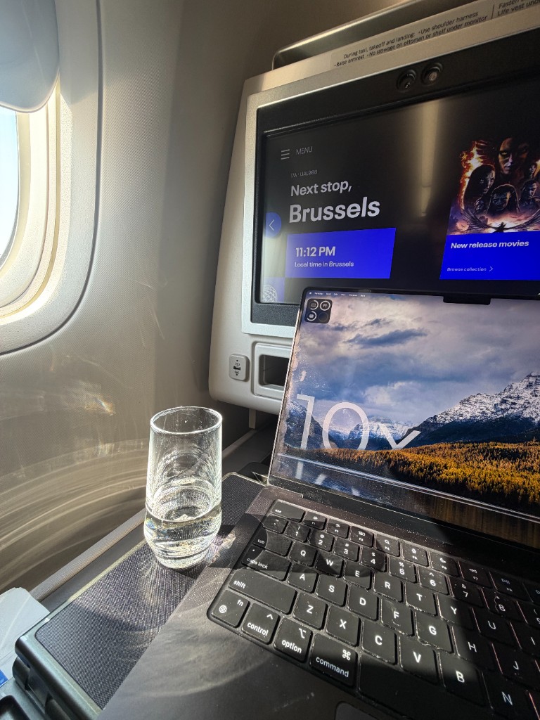 A 10x laptop open on an airplane tray table with the seatback screen showing next stop Brussels.